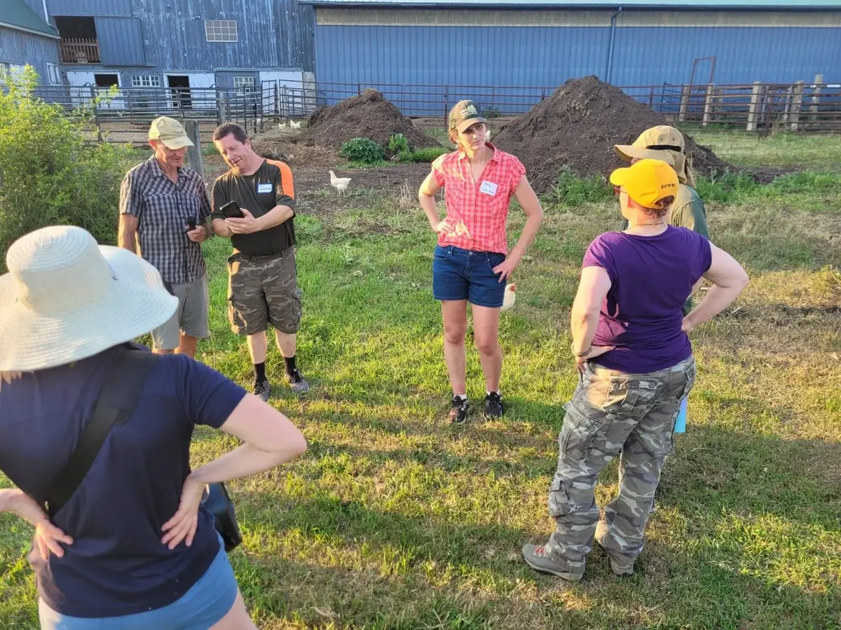 Visitors in sun hats stand on the grass at Good Trouble Grove while the farmers talk them through the farm
