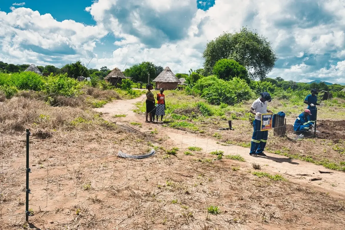 A new fence line runs beside a sandy path toward thatched huts in Masoka while workers check equipment