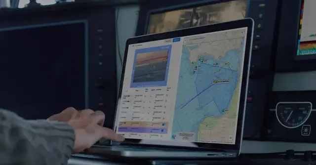 A hand on a laptop aboard a boat, showing the M2 vessel-tracking map beside a live camera feed