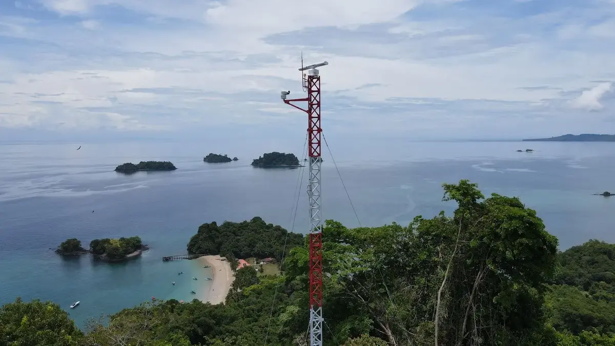 A red-and-white M2 radar and camera tower on a forested hill above a bay and small islands