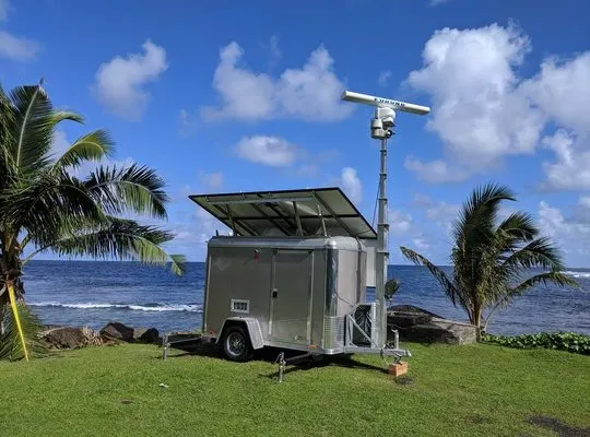 A solar-powered M2 trailer with a radar mast parked on grass by the sea, between palm trees