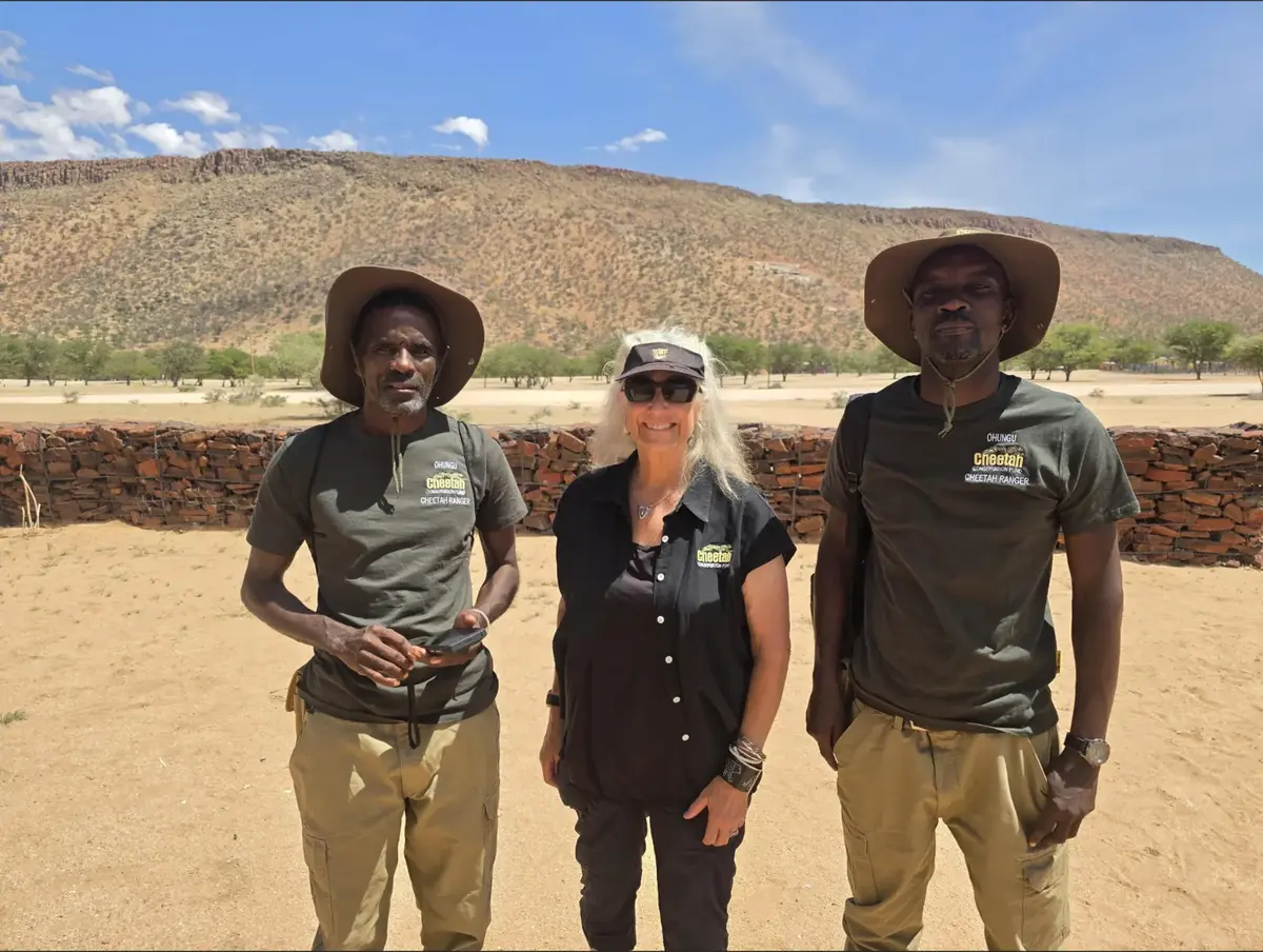 Dr. Laurie Marker stands between two Cheetah Rangers in front of a stone kraal and a flat-topped hill